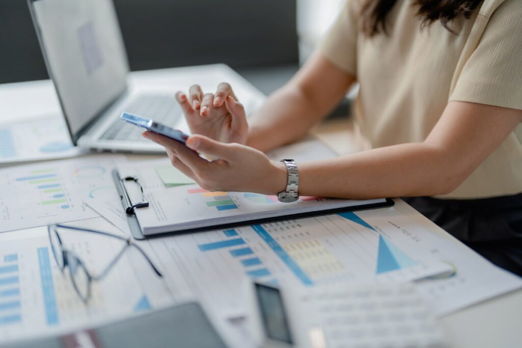 Pessoa analisando gráficos financeiros no celular, com papéis de contas a pagar e receber ao lado.