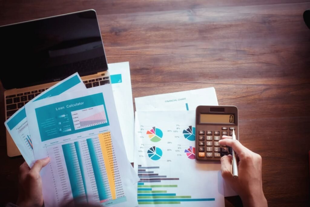 Mãos calculando tarifas em uma mesa com gráficos e laptop, representando tarifação telefônica.