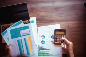 Mãos calculando tarifas em uma mesa com gráficos e laptop, representando tarifação telefônica.