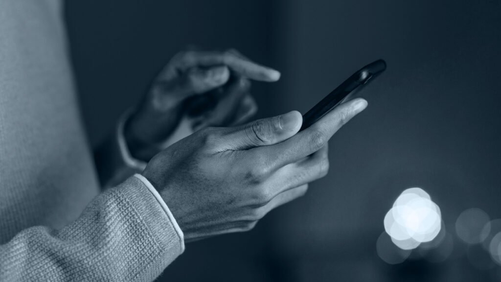 Pessoa com camisa de manga cumprida, segurando um celular com a mão esquerda e digitando com a direita.