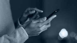 Pessoa com camisa de manga cumprida, segurando um celular com a mão esquerda e digitando com a direita.