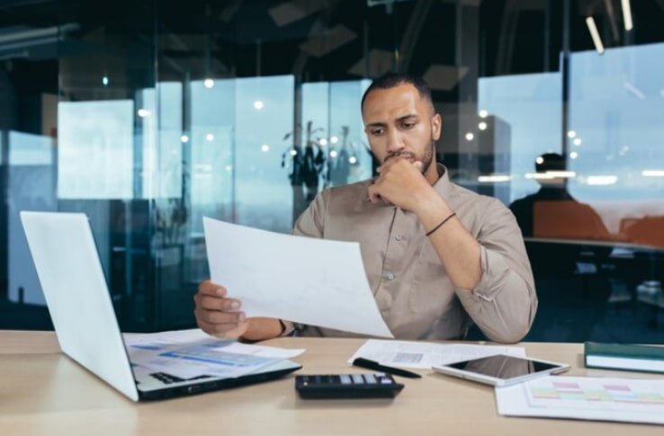 Homem sentado em frente a notebook e com papel na mão pensando sobre custo padrão e custo real
