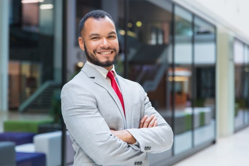 Homem negro de roupa social e gravata vermelha, com os braços cruzados e sorridente.