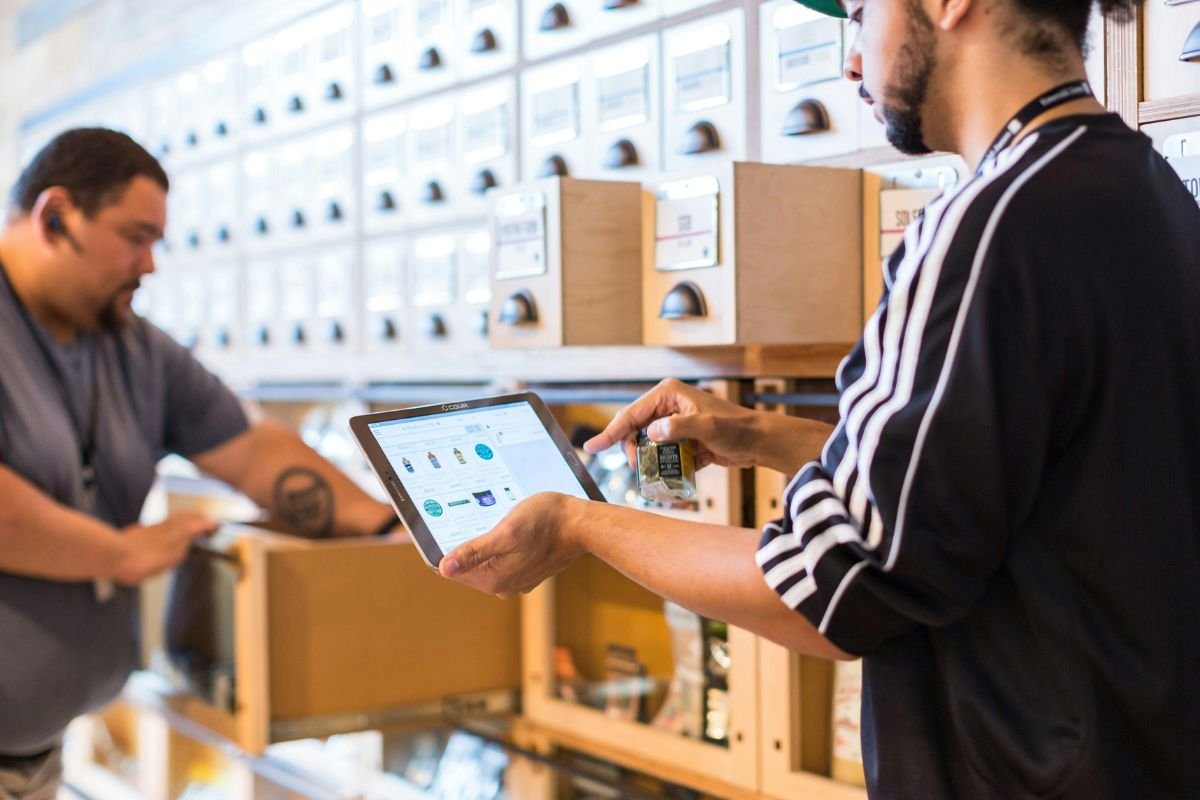 Dois homens no setor de estoque da empresa. Um deles segura o tablet com as informações dos produtos inventariados; o outro manuseia as caixas com os produtos.