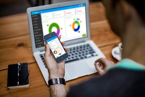 Homem de roupa social, observando gráficos e dados em tela de celular e do notebook.