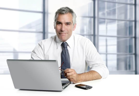 Homem de roupa social branca e grava azul, usando um notebook cinza sobre a mesa.