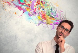 Homem de roupa social, óculos e gravata preta, pensando, com uma parede branca suja de tinta atrás.