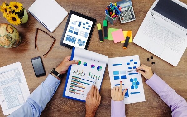 Mãos de duas pessoas segurando folhas com gráficos. Na mesa, também há um tablet com gráficos em sua tela, uma calculadora, um notebook, um celular, alguns post-its, um caderno e dois vasos de flores.