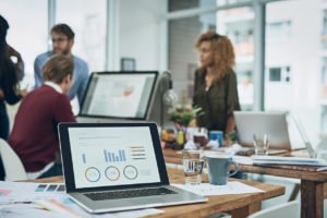 Pessoas em um escritório, com u notebook em cima de mesa de madeira, com gráficos, ao lado de um copo de água.